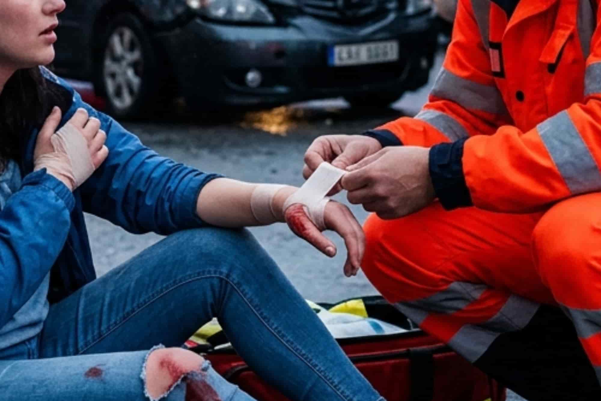 A first responder in an orange uniform bandages the arm of an injured woman sitting on a street after a car accident.